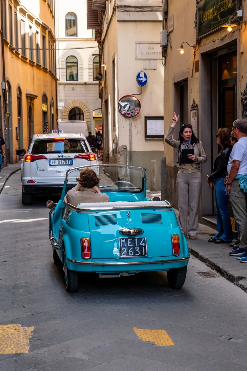 A vintage blue convertible driving along a narrow historic street in Florence.