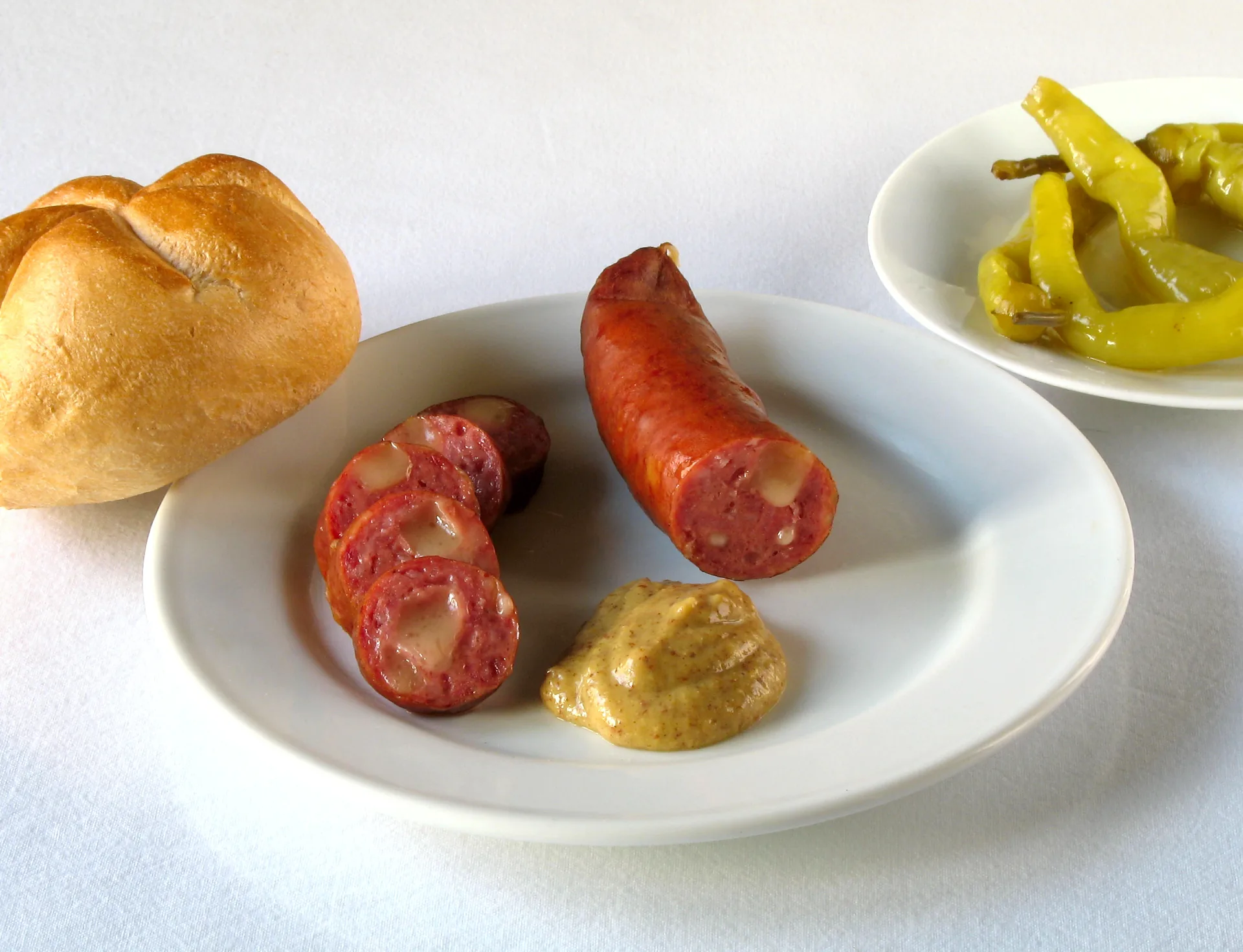 A Käsekrainer sausage served with mustard, a bread roll and pickled peppers.