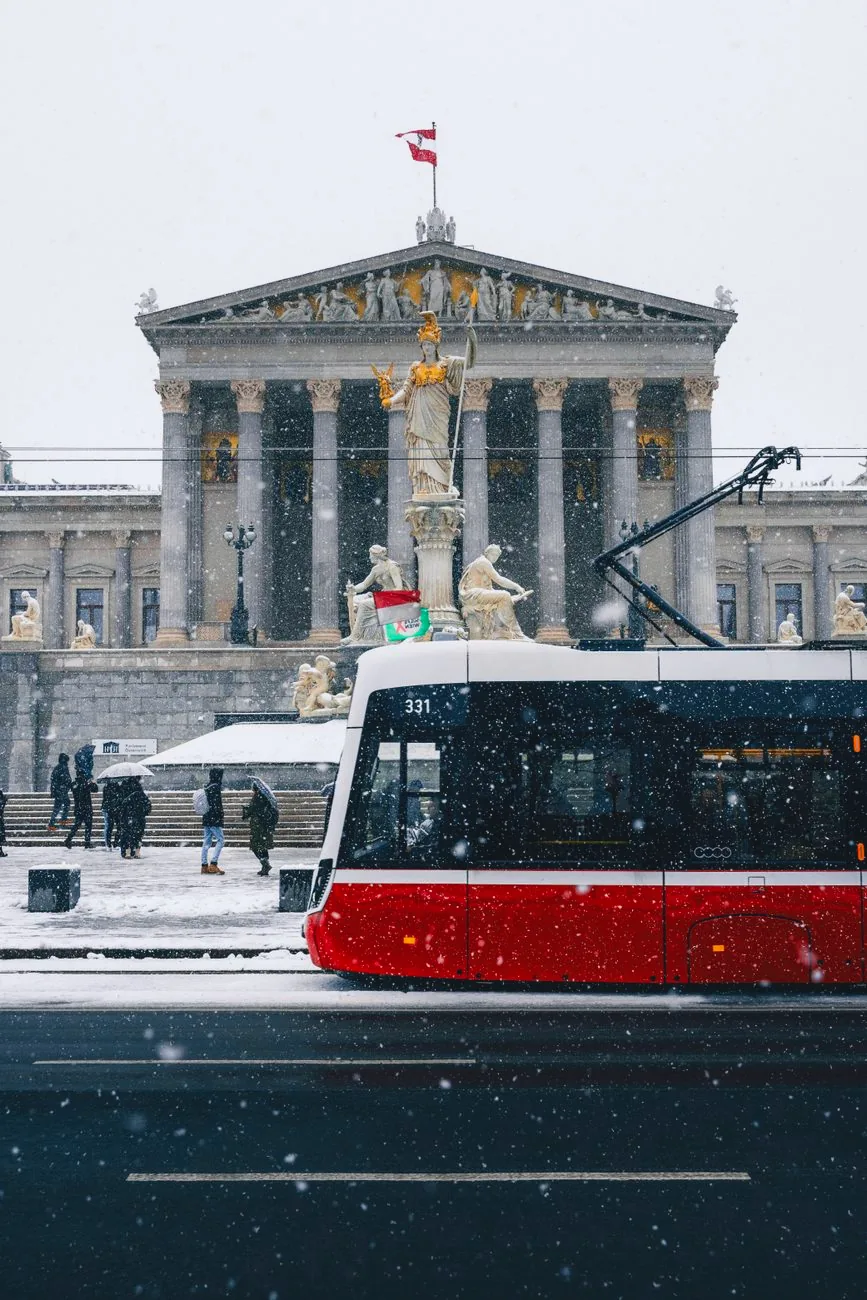 A red tram passes the Austrian Parliament in Vienna on a snowy day.