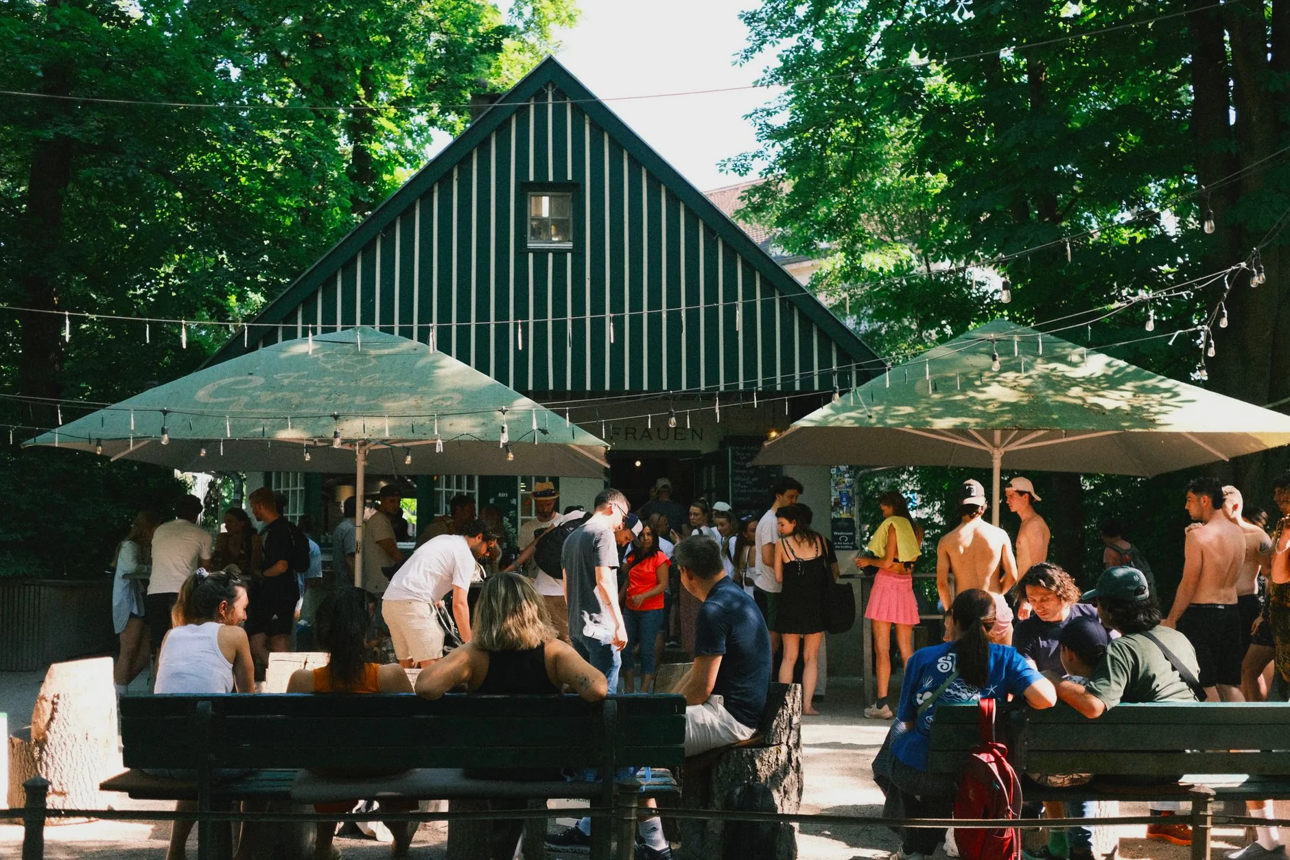 People sharing drinks at a busy Munich beer garden, seated beneath trees at communal tables.