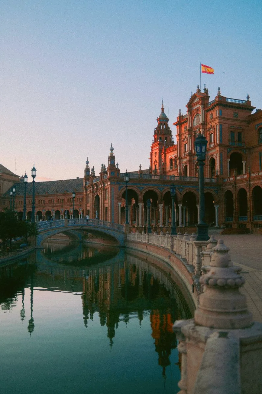 Plaza de España’s curved brick buildings reflected in the canal, with a bridge crossing the water.