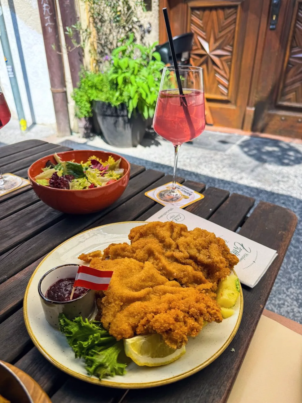 A breaded schnitzel served with salad and a drink at an outdoor table.