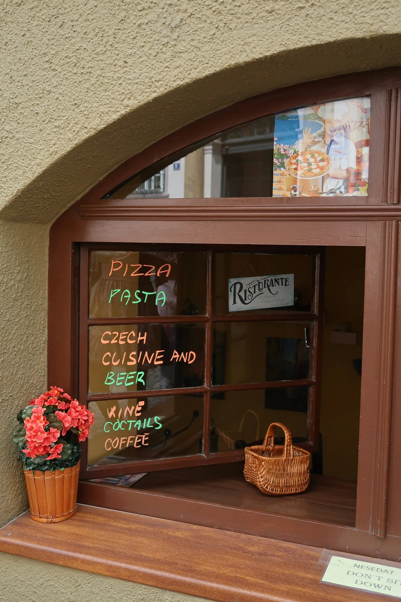 A restaurant window in Prague with a menu, flowers and a small basket.