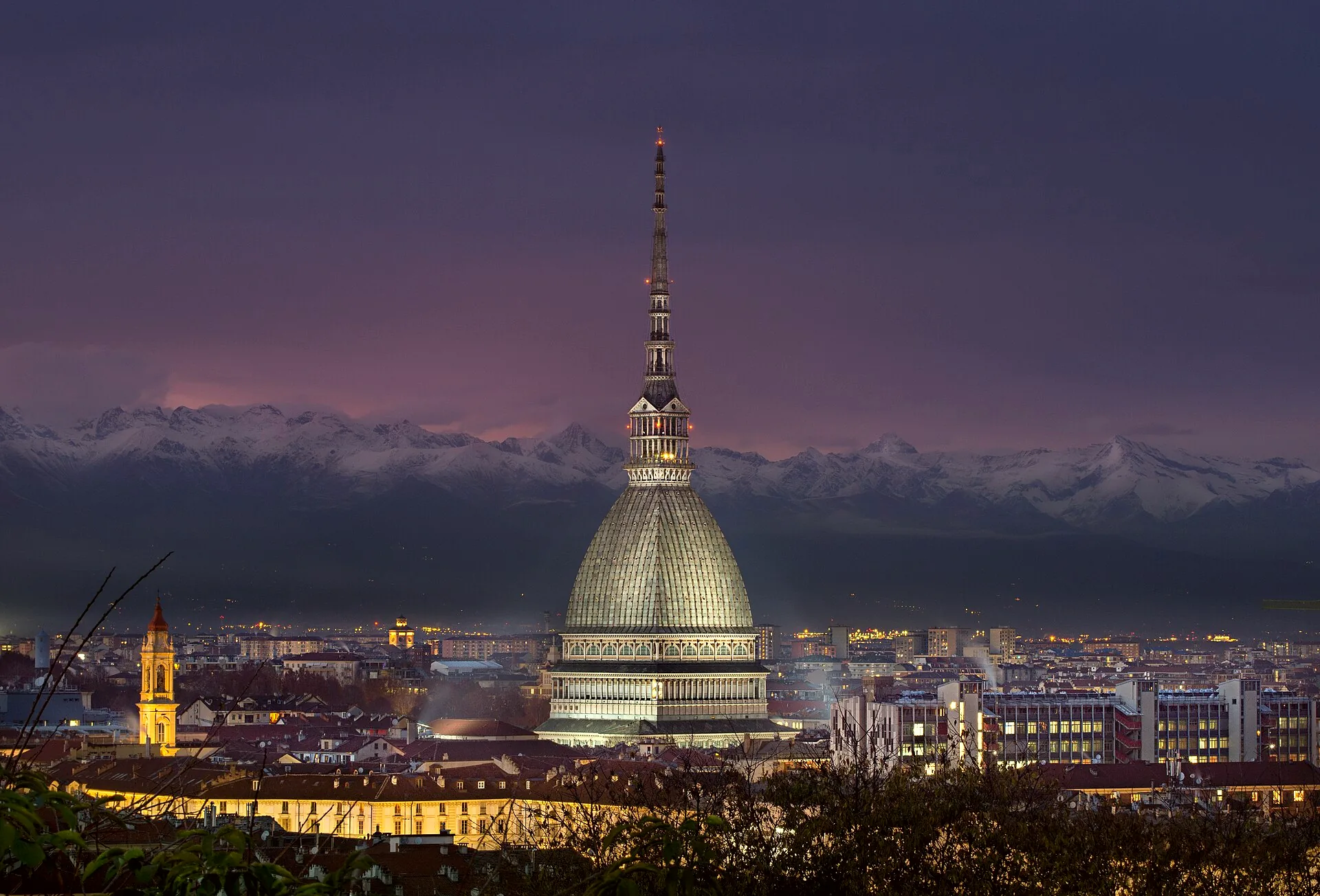The Mole Antonelliana illuminated at night, with the Alps in the background.