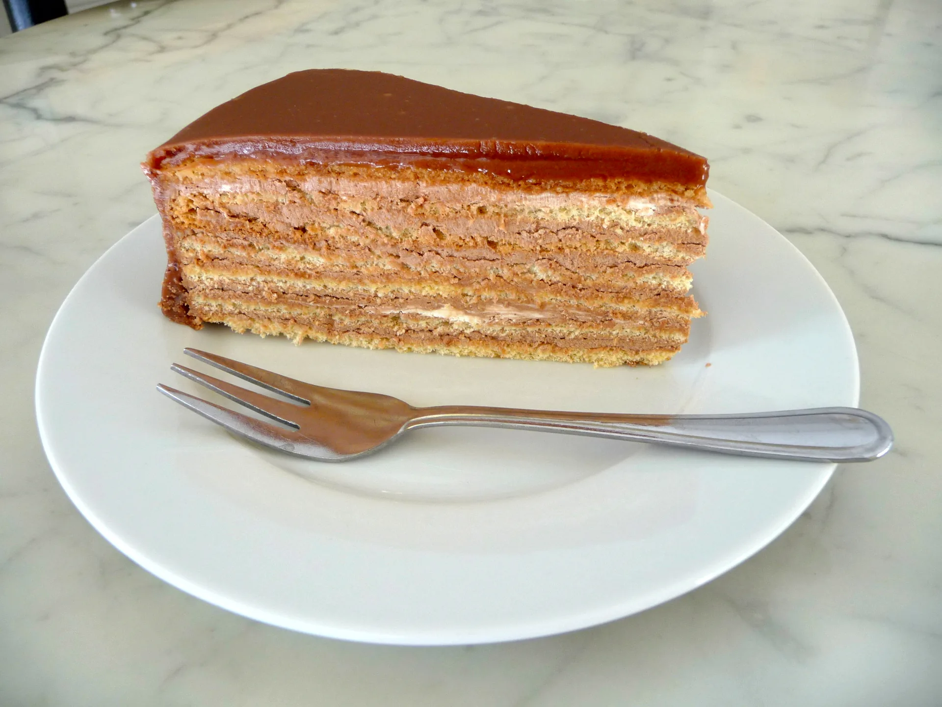 A slice of Prinzregententorte, with layers of sponge and chocolate buttercream beneath a dark glaze.