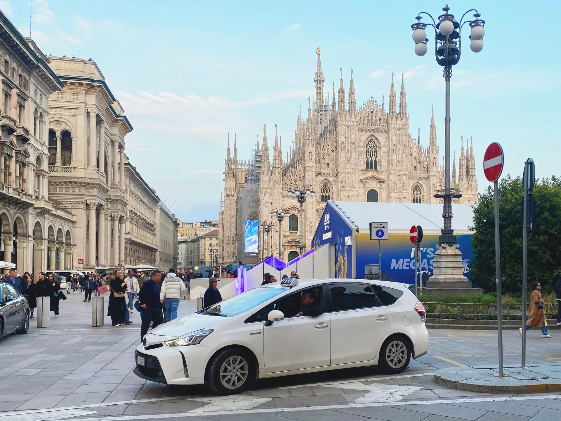 A white Toyota Prius+ taxi with a rooftop sign in front of Milan Cathedral.