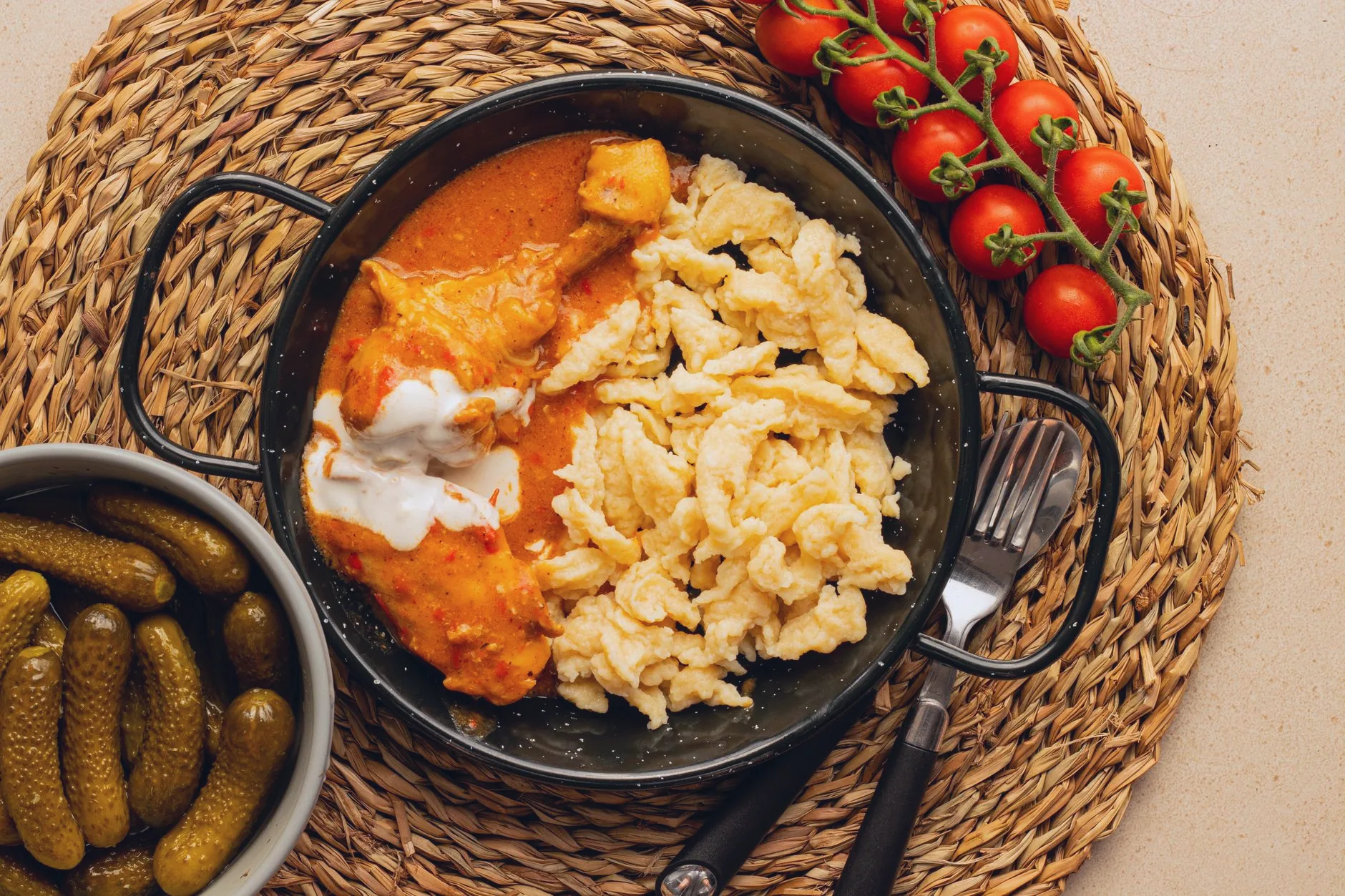 Chicken paprikash with nokedli, tomatoes and pickles on a woven placemat.
