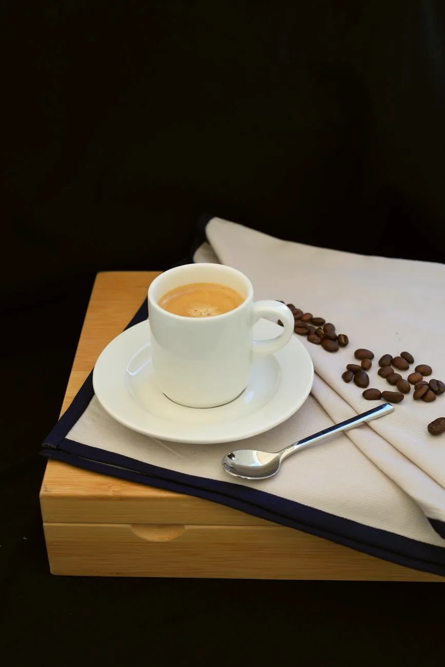 A white cup of espresso on a saucer, with coffee beans and a spoon on a wooden surface.