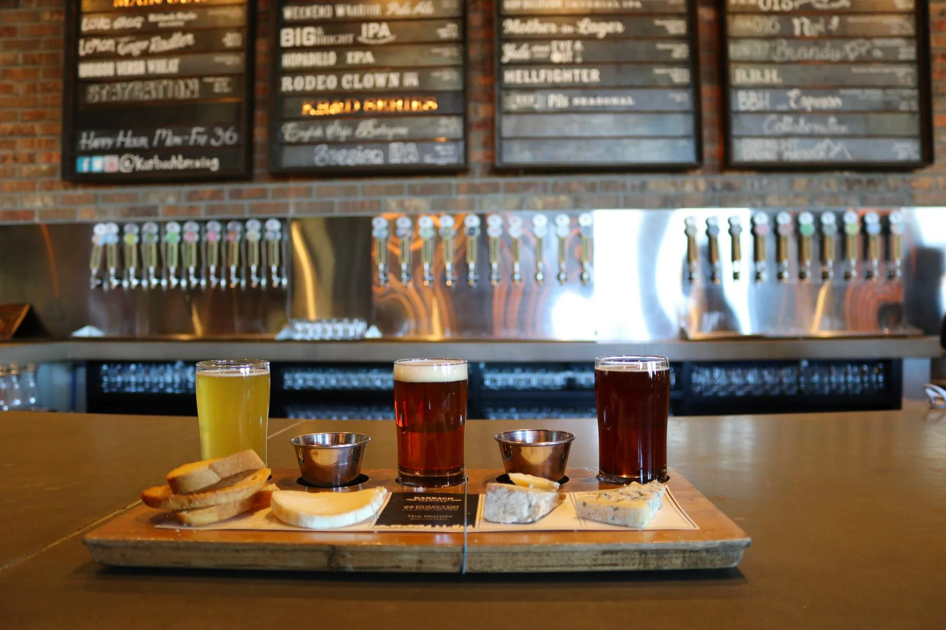 Several glasses of craft beer beside cheese and bread on a bar counter.