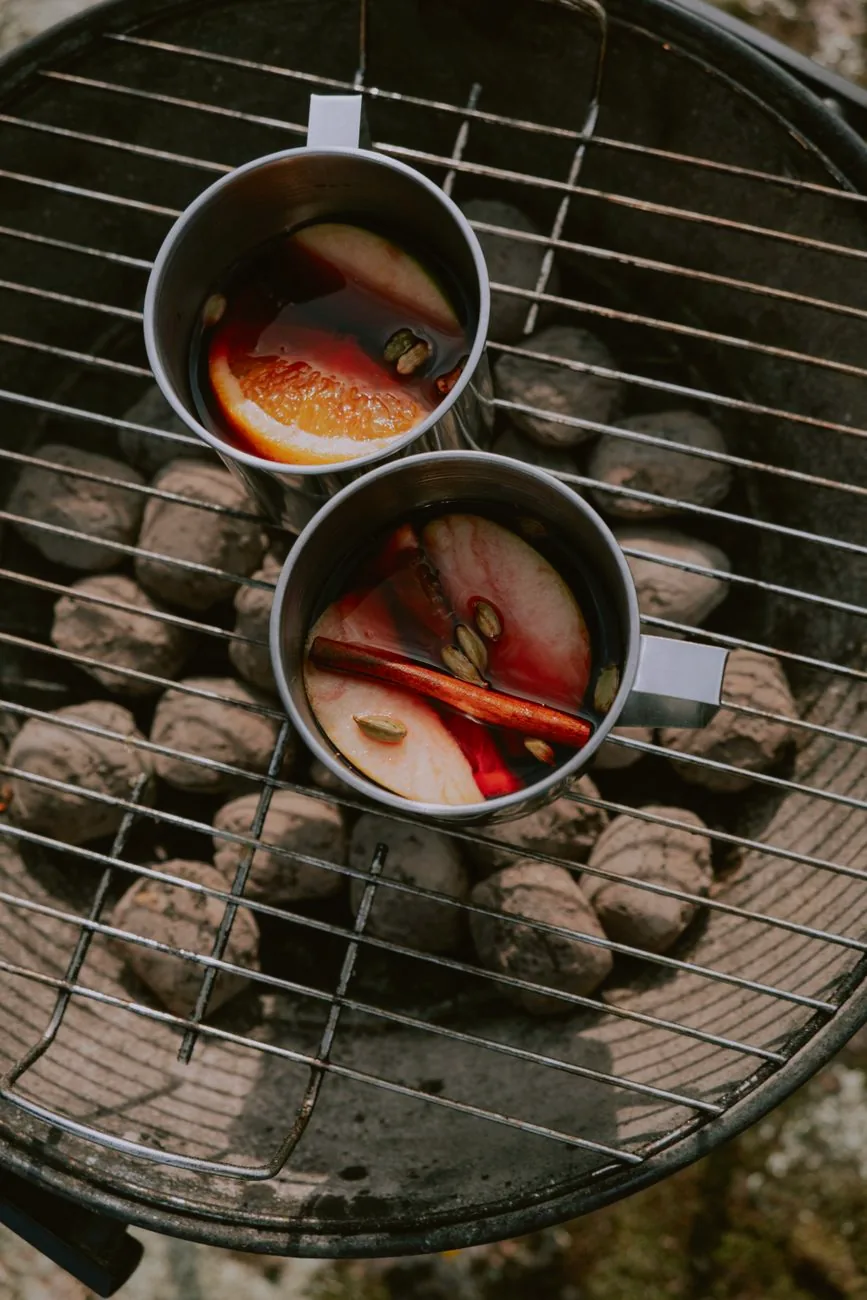Cups of mulled wine beside aromatic spices on a charcoal grill.