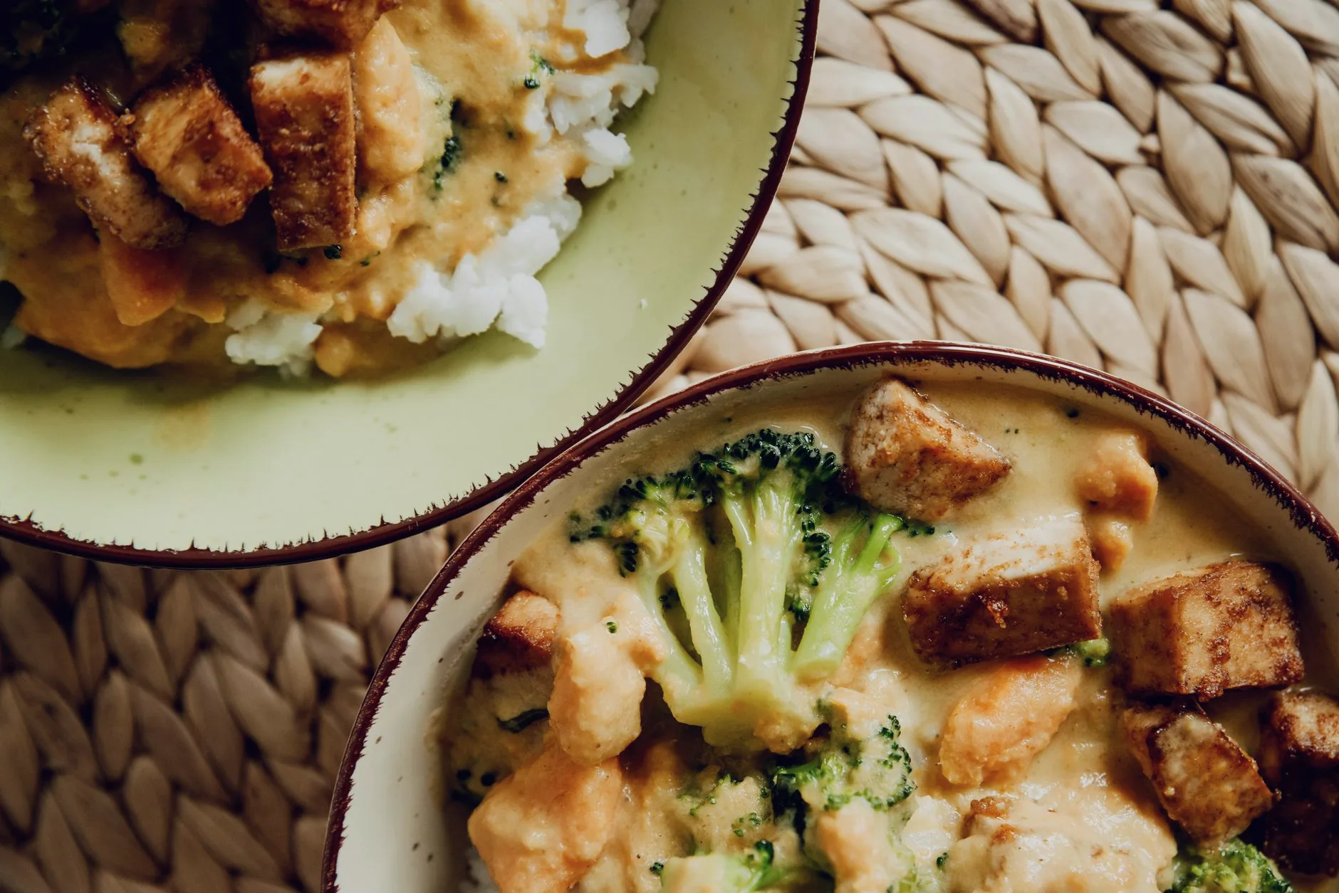 A bowl of tofu and broccoli with creamy sauce on a woven mat.