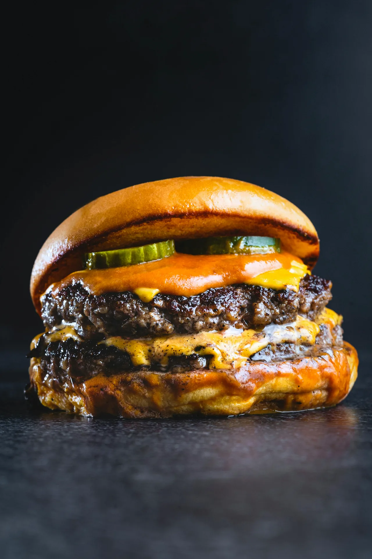 A double cheeseburger with pickles on a dark surface.